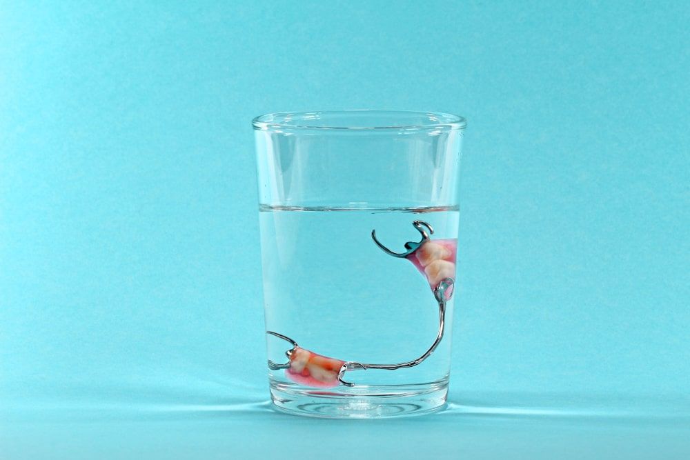 Partial denture soaking in a glass of water against a light blue background